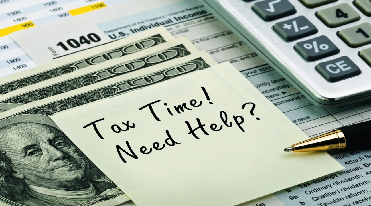 A sticky note reading Tax Time! Need Help? rests on U.S. $100 bills, with IRS 1040 tax forms, a pen, and a calculator partially visible in the background.