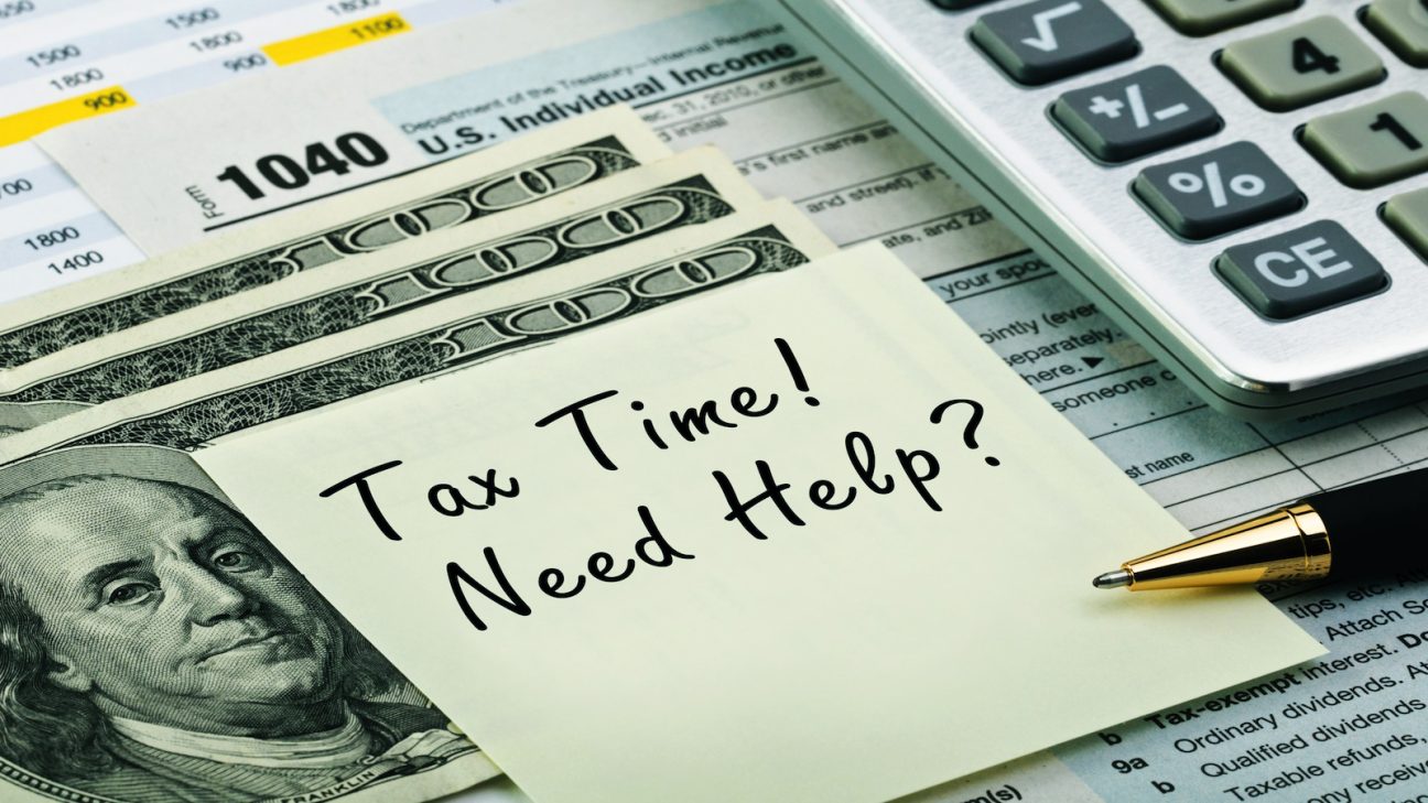 A sticky note reading Tax Time! Need Help? rests on U.S. $100 bills, with IRS 1040 tax forms, a pen, and a calculator partially visible in the background.